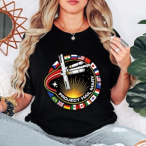 May include: A black t-shirt featuring a circular graphic with a space station illustration, a bright yellow sunburst, and the text "PROJECT HAIL MARY". The border of the circle is decorated with various international flags arranged in a ring.