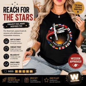 May include: A black t-shirt featuring a circular graphic with a space shuttle illustration, the text "PROJECT HAIL MARY," and a border of international flags. The design includes a sunburst effect and a red orbital path on a dark background.