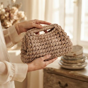 May include: A beige handbag crafted from thick, chunky yarn in a dense crochet stitch pattern. The bag features a structured, rectangular frame with an integrated handle opening at the top, showcasing a textured, knotted surface design.