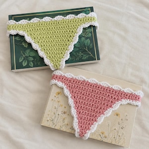 May include: Two triangular crochet bandanas, one in light green and one in dusty pink, each featuring a white scalloped edge border. The green bandana is placed on a dark green book, and the pink bandana is placed on a cream-colored book.