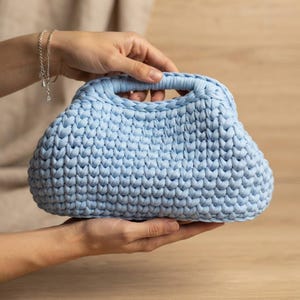 Può includere: Una borsa a mano azzurra realizzata con filato spesso e voluminoso in un motivo a uncinetto. La borsa presenta una forma arrotondata e strutturata con un manico integrato nella parte superiore, mostrando una trama densa e uniforme.