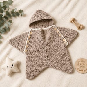 May include: A star-shaped crochet baby bunting bag in a light brown color with a hood. The garment features a white trim along the front opening and is secured with a series of small wooden buttons. The texture is consistent throughout the piece.