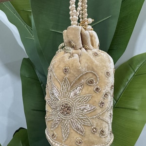 May include: A beige velvet drawstring pouch bag featuring intricate gold beadwork in a floral pattern. The bag is accented with small clear beads and includes a long handle made of multiple strands of cream-coloured faux pearls for carrying.