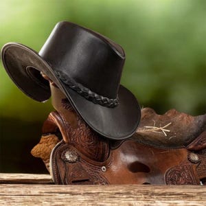 May include: A dark brown leather cowboy hat with a braided leather hatband resting on a brown leather saddle featuring intricate embossed floral patterns and silver-toned metal hardware accents on the side of the saddle structure.