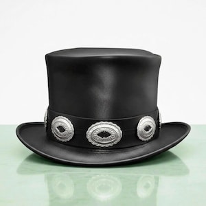 May include: A black top hat featuring a wide band around the base decorated with a series of oval-shaped silver-toned metal conchos. The hat has a flat crown and a curved brim, constructed with a smooth, matte finish throughout the exterior.
