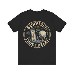 Può includere: Una t-shirt nera con una grafica circolare sul retro. Il design include un termometro sorridente, una pallina da golf su un tee e dei fiocchi di neve. Il testo SURVIVED THE FROST DELAY è disposto in cerchio attorno all'immagine centrale.