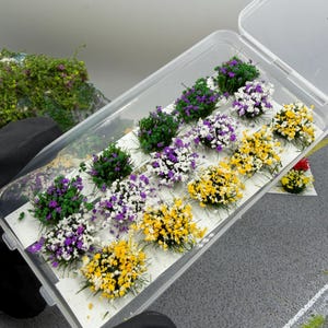 Peut inclure: Une boîte de rangement en plastique transparent contenant plusieurs touffes de fleurs miniatures de différentes couleurs, notamment jaune, violet, blanc et vert. Ces petits groupes texturés sont disposés en rangées sur un support blanc.