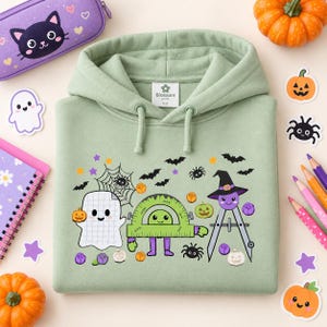 May include: A sage green hooded sweatshirt featuring a graphic print of a grid-patterned ghost, a green protractor, and a purple compass wearing a witch hat, surrounded by small bats, spiders, spiderwebs, pumpkins, and purple flowers.