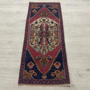 May include: A rectangular area rug featuring a central diamond-shaped medallion in cream, surrounded by a deep red field and a dark navy blue border. The design includes intricate geometric patterns in shades of green, orange, and dark blue.