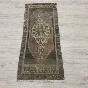 May include: A rectangular area rug featuring a central diamond-shaped medallion with geometric floral motifs in shades of muted purple, dark brown, and cream. The rug has a wide decorative border with repeating patterns and short white fringe ends.