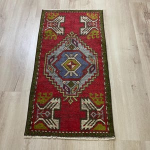 May include: A rectangular area rug featuring a vibrant red background with a central blue diamond motif containing geometric patterns in green, white, and purple. The rug is framed by a dark green border and includes white fringe at both ends.