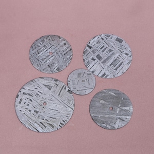 May include: Five circular, flat metal discs featuring a distinct, intersecting crystalline Widmanstätten pattern in shades of grey and silver. Four of the discs have a small, centered circular hole, while the smallest disc is solid.