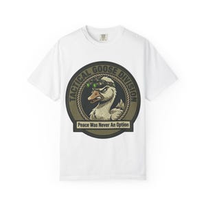 Peut inclure: Un t-shirt blanc avec un motif circulaire représentant une oie portant des lunettes de vision nocturne. Le texte indique TACTICAL GOOSE DIVISION et Peace Was Never An Option. Le design utilise des tons vert olive, noir et blanc.