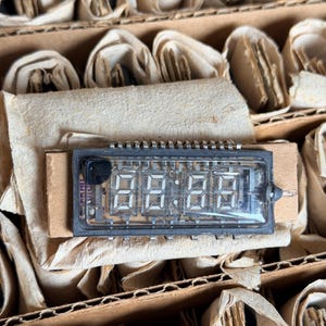 May include: A rectangular vacuum fluorescent display module featuring a clear glass casing with a black frame. The internal component displays a four-digit numerical sequence with a colon in the center, mounted on a piece of light brown cardboard.