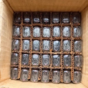 May include: A collection of thirty-five glass nixie tubes arranged in a grid within a cardboard box. Each rectangular tube contains thin metal wire filaments shaped into numerical digits and a fine wire mesh screen, all encased in clear glass.
