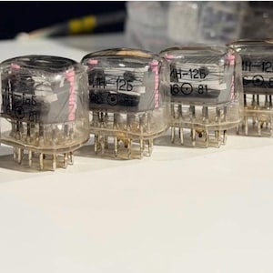 May include: A row of six clear glass nixie tubes with internal metal components and pink accents. Each tube features the printed text "ИН-12Б" and "06 81" on the front. The electronic components are visible through the transparent glass housing.
