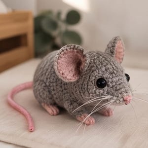 May include: A small, handcrafted crochet mouse toy made with grey yarn. It features large, rounded ears with pink inner detailing, a small pink nose, black plastic safety eyes, and long white whiskers. A long, thin pink tail extends from the back.
