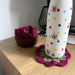 May include: A white metal water bottle with a pattern of colorful polka dots and bumblebees, resting on a round, flower-shaped crochet coaster in magenta and green, next to a small, brown, bowl-shaped crochet item with magenta petals.