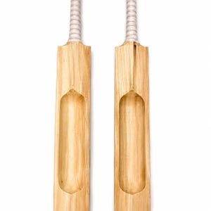 May include: Two light-colored wooden cricket bats positioned vertically side-by-side. Each bat features a smooth, natural wood grain finish, a long central concave groove on the front face, and a handle wrapped in white and red corded grip tape.