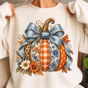 Pode incluir: Uma camisola de gola redonda cor de creme com um grande gráfico de uma abóbora adornada com um laço azul. A abóbora está dividida em secções verticais com padrão de leopardo, xadrez laranja, flores azuis e bolinhas cor de laranja.