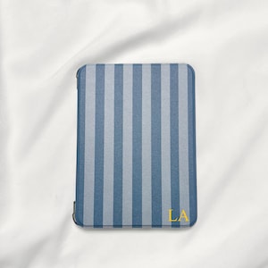 May include: A rectangular e-reader case featuring a vertical striped pattern in alternating shades of blue and light gray. The bottom right corner of the case displays the yellow capital letters LA, which are printed in a simple serif font.