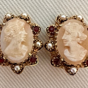 Könnte beinhalten: Ein Paar Vintage-Ohrringe mit Kamee. Jeder Ohrring zeigt ein geschnitztes Profil in einem ovalen Rahmen. Die goldfarbenen Rahmen sind mit kleinen Perlen und roten Edelsteinen verziert. Die Ohrringe sind vor einem neutralen Hintergrund platziert.