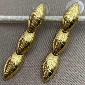 Könnte beinhalten: Ein Paar goldfarbene Ohrringe mit geometrischem Design. Jeder Ohrring besteht aus drei miteinander verbundenen ovalen Formen mit einer strukturierten Oberfläche.