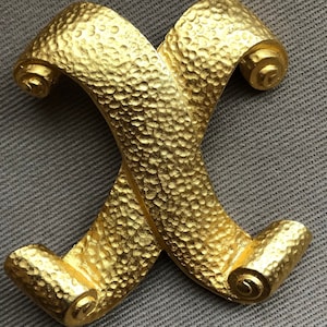 May include: A gold-colored metal brooch in the shape of two scrolls that are intertwined to form an 'X'. The scrolls have a textured surface.