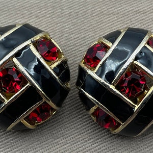 May include: A pair of black and gold tone clip-on earrings with red rhinestone accents. The earrings are round and have a woven design.