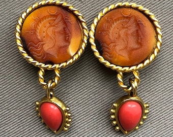 May include: A pair of gold tone earrings with a cameo-style pendant featuring a woman's profile in relief. The cameo is set in a round, gold-tone frame with a rope-like border. The pendant is attached to a gold-tone chain that ends in a teardrop-shaped coral stone.