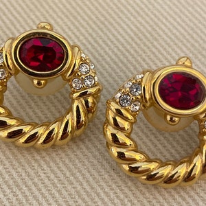 Puede incluir: Un par de pendientes de tono dorado con un diseño de cuerda trenzada. Cada pendiente presenta una piedra grande de color rojo ovalada rodeada de piedras más pequeñas de color claro.