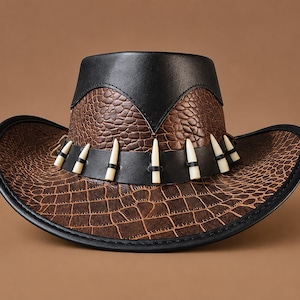 Peut inclure: Un chapeau marron et noir avec un motif texturé en peau de crocodile sur la calotte et les bords. Le chapeau comprend une bande noire ornée d'une série d'accents blancs pointus en forme de dents entourant la base de la calotte.