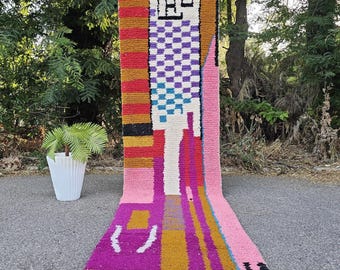 May include: A long, rectangular runner rug featuring a vibrant, multi-colored geometric pattern. The design includes blocks of pink, mustard yellow, purple, and black, with a central section of white and blue checkered squares and white fringe.