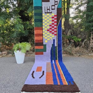 May include: A long, rectangular hand-tufted rug featuring a vibrant geometric pattern with blocks of yellow, red, blue, orange, green, and purple. The rug includes a checkered section, abstract shapes, and a dark brown fringe at the bottom edge.