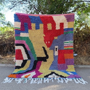 May include: A rectangular area rug featuring a high-pile texture and a vibrant, multicolored geometric pattern. The design includes blocks of red, yellow, blue, green, purple, orange, black, and cream, finished with white fringe at the bottom.