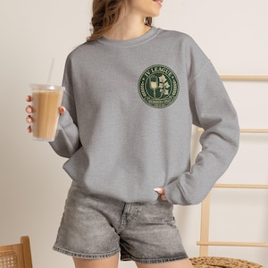 May include: A heather gray crewneck sweatshirt featuring a circular green emblem on the left chest. The emblem displays an intravenous drip bag, ivy leaves, and the text IV LEAGUE MEMBERS ONLY. The sweatshirt has long sleeves and a ribbed neckline.