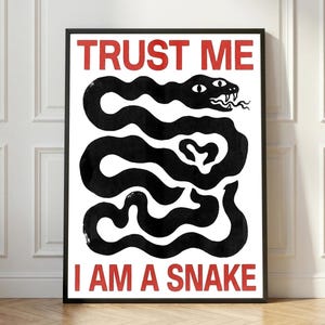 Op de afbeelding: Een rechthoekige wandkunstprint in een zwarte lijst met een witte achtergrond en een grote, gestileerde zwarte slangillustratie in het midden. De tekst TRUST ME staat in het rood bovenaan en I AM A SNAKE staat in het rood onderaan.