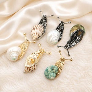 Puede incluir: Una colección de seis broches con forma de caracol que presentan cuerpos de metal texturizado en tonos dorados y plateados. Cada caracol tiene una concha espiral natural, mostrando varios patrones y colores incluyendo blanco, marrón y verde.