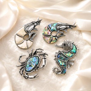 May include: Four silver-toned metal brooches shaped like sea creatures. One shrimp features white shell segments, another shrimp has iridescent abalone shell inlays, a crab displays an abalone shell center, and a seahorse features abalone shell.