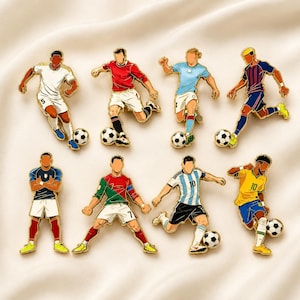 Può includere: Una collezione di otto spille smaltate raffiguranti calciatori con diverse divise di squadra, ognuno in una posa dinamica con un pallone. I giocatori indossano maglie nei colori bianco, rosso, azzurro, blu navy, blu scuro, verde e giallo.