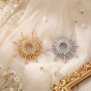 Puede incluir: Dos broches con forma de sol, uno en tono dorado y otro en tono plateado, cada uno con un anillo circular central incrustado con pequeños cristales transparentes y rayos metálicos ondulados que irradian desde el centro.