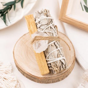 May include: A bundle of dried white sage wrapped in white string, a single rectangular stick of light brown palo santo wood, and a clear, jagged quartz crystal, all tied together with brown twine and resting on a circular wooden slice.