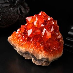 May include: A cluster of vibrant orange citrine crystals with sharp, multifaceted points. The upper section features translucent, deep amber-colored crystalline structures, while the base consists of a solid, opaque, light-colored rocky matrix.