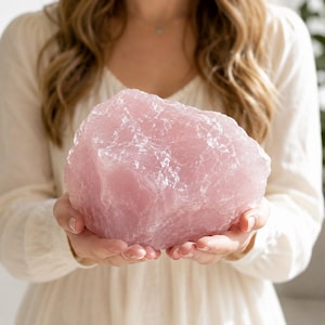 Può includere: Un grande pezzo grezzo di cristallo di quarzo rosa traslucido e pallido con una consistenza superficiale irregolare e frastagliata, caratterizzato da fratture cristalline naturali. La pietra mostra varie tonalità di rosa tenue e bianco.