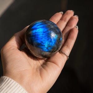 May include: A polished labradorite sphere featuring a dark grey base with a vibrant, iridescent blue flash across its surface. The stone is spherical in shape, displaying natural mineral inclusions and a smooth, reflective texture throughout.