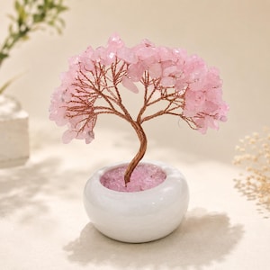 Puede incluir: Una escultura de árbol decorativa con un tronco y ramas de alambre de cobre trenzado, adornados con numerosas pequeñas virutas de cuarzo rosa. El árbol está colocado en una maceta de cerámica blanca, redonda y lisa, llena de cristales rosas.