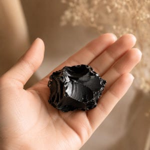 Puede incluir: Una pieza bruta y sin pulir de piedra obsidiana negra con una textura brillante similar al vidrio y bordes afilados e irregulares. La piedra presenta un color negro profundo y sólido con una superficie reflectante que capta la luz.