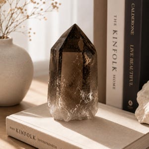 Puede incluir: Una punta de cristal de cuarzo ahumado pulido con un tono marrón oscuro translúcido y fracturas internas naturales. El cristal presenta una forma geométrica hexagonal con una parte superior puntiaguda, mostrando un degradado de color.