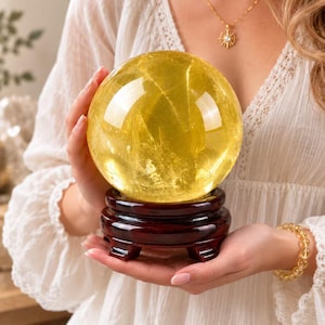 May include: A large, spherical, translucent yellow crystal ball with visible internal fractures and inclusions, resting on a dark, polished, circular wooden stand with four small feet, showcasing a smooth, reflective surface and vibrant golden hue.