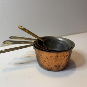 May include: A set of four nested measuring cups with a weathered copper exterior and tarnished brass handles. Each cup features a long, flat handle with a small hole at the end for hanging. The metal surfaces show signs of age, patina, and wear.